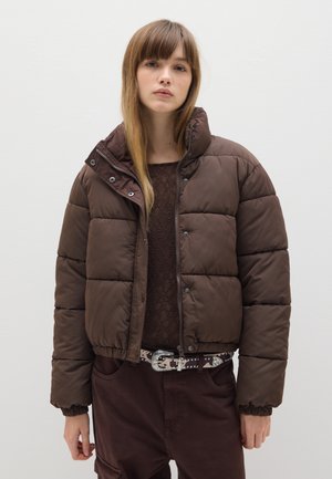 Braune Pufferjacke mit cropped Design, gestepptem Muster, Druckknöpfen und elastischen Bündchen, kombiniert mit einem dunklen Spitzentop und braunen Hosen.
