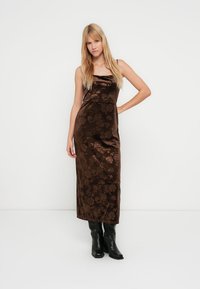 Brązowa aksamitna sukienka maxi w kwiatowy wzór, dopasowana w biuście, o prostej sylwetce i cienkich ramiączkach, noszona z czarnymi wysokimi kozakami za kolano.