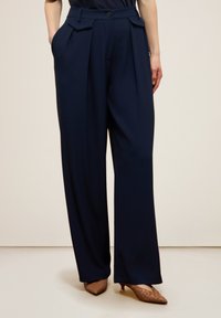 Navyblauwe wijde broek met een hoge taille, voorzien van voorplooien, knoopsluiting en zijzakken. Vloeiend stoffen textuur, gekleed ontwerp.