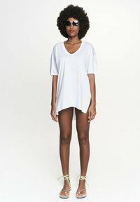 Biała, oversize'owa koszulka z dekoltem w serek, krótkimi rękawami i zaokrąglonym dołem. Noszona z zielonymi sandałami z wiązaniami na kostkach oraz dużymi czarnymi okularami przeciwsłonecznymi.