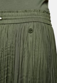 Close-up van een olijfgroene geplooide rok met elastische tailleband en een klein rond logo-badge aan de voorkant.