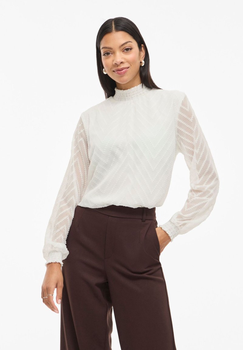 Weiße Bluse mit einem hohen Rüschenausschnitt und langen Ärmeln. Sie hat ein Zickzackmuster und einen durchsichtigen, strukturierten Stoff. Kombiniert mit dunklen Hosen.