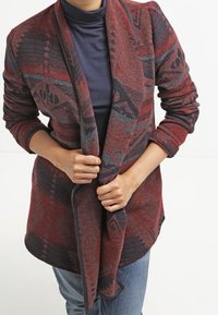 Personne portant un cardigan ouvert à motifs rouge et noir sur un col roulé bleu marine et un jean bleu, ajustant le cardigan avec les deux mains.