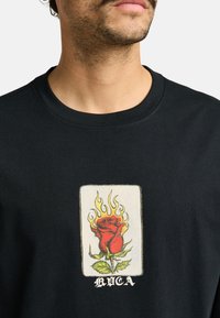 Black cotton t-shirt featuring a flame-accented red rose graphic in the center, with "RICA" text below in white. Classic crew neck design.