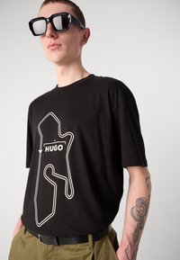 Melnas kokvilnas T-krekls ar baltu grafisko dizainu, kurā redzams teksts "HUGO". Modelis valkā pārmērīgi lielas melnas saulesbrilles un melnu jostu.