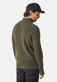 Dark green, ribbed sweater with a high collar and long sleeves. Features a textured fabric and a straight hem. Worn with a black cap.