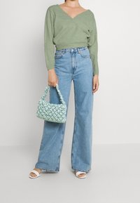 Kobieta ubrana w zielony sweter w serek i spodnie z szerokimi nogawkami, trzymająca jasnoniebieską plecioną torebkę, stojąca w białych sandałach na obcasie.