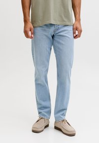 Bărbat purtând blugi bleu deschis, pantofi bej din piele întoarsă și un tricou verde măsliniu, stând într-un fundal alb simplu.