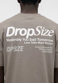 Personne portant un t-shirt beige avec texte blanc "DropSize", phrases motivantes et "Limited Release" imprimées dans le dos.