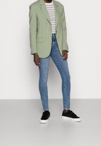 Grüner Blazer, gestreifes T-Shirt, schmal geschnittene hellblaue Jeans und schwarze Sneaker. Der Blazer hat ein Revers mit Schlitz und einen einzelnen Knopfverschluss.