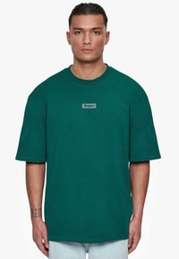 Camiseta oversized de color verde azulado con mangas cortas, escote redondo y una etiqueta cosida que dice "Dropsize" en el pecho, fabricada en tejido de algodón.