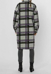 Abrigo a cuadros en tonos gris, negro y púrpura, hecho de una tela suave. Presenta un corte largo y oversize con puños abotonados. Llevado sobre pantalones vaqueros ajustados.