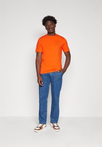 Oranges Baumwoll-T-Shirt mit kurzen Ärmeln und einem kleinen Logo auf der Brust, kombiniert mit blauen Jeans und mehrfarbigen Sneakers.