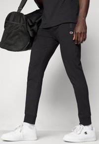 Zwarte joggingsbroek met een slim fit, elastische boorden en zijnaad. Gecombineerd met witte sneakers en een zwarte sporttas.