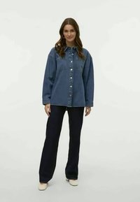 Tummansinisestä denim-painepaitasta, jossa on nappisuljenta, kaksi rintataskua ja ylisuurta muotoa; yhdistettynä leveälahkeisiin mustiin housuihin ja vaaleisiin kenkiin.