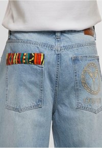 Helle blaue Denim-Jeans mit zwei Gesäßtaschen, von denen eine mit buntem gestreiftem Stoff und die andere mit einem gestickten runden Emblem und Text versehen ist.
