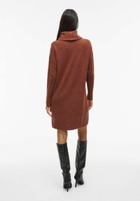 Obleka s turtleneck iz rjave rje, z pleteno teksturo, dolgimi rokavi in ribanim robom, v kombinaciji s črnimi visokimi škornji s špicami do kolen.