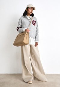 Pull en tricot gris avec des rayures bordeaux et logo 'G', pantalon ample beige, et sac à épaule tan sur fond uni.