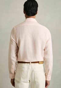 Cămașă din in roz deschis, cu mâneci lungi și mansete cu nasturi, văzută din spate. Purtați-o cu pantaloni albi și o curea maro.