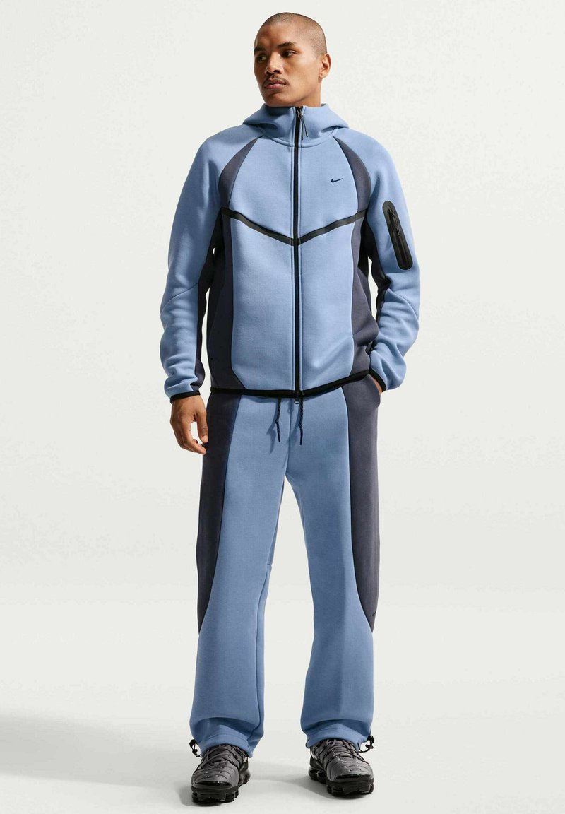 Homme debout portant une veste à capuche zippée bleu et gris foncé avec un pantalon assorti et des chaussures de sport noires, les mains dans les poches, regardant à gauche.