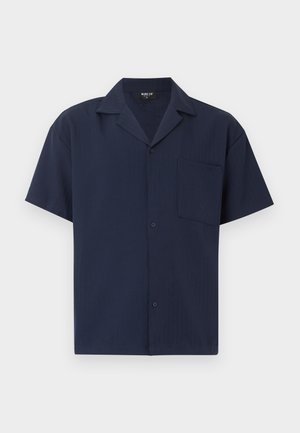 Chemise bleu marine à manches courtes avec boutons, col pointu et une unique poche poitrine, posée à plat sur un fond blanc.