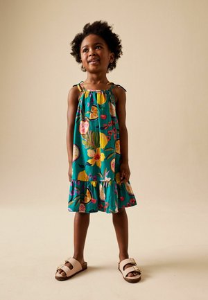 Junges Mädchen mit lockigem Haar, das ein ärmelloses, türkisfarbenes Kleid mit Obst- und Blumenmuster trägt und barfuß vor beigem Hintergrund steht.