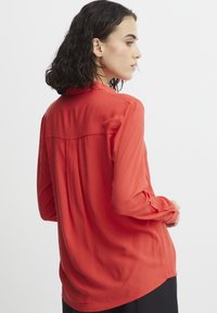 Blusa rossa a maniche lunghe in tessuto morbido. Presenta un yoke posteriore, pieghe sottili e maniche con polsini. L'orlo è arrotondato.