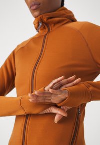 Orange zip-up jacka med hög krage, med en mjuk textur och tumhål vid ärmsluten, kompletterad med en sidofickdragkedja.