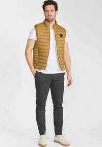 Gevoerde tan gilet met hoge kraag en zwart embleem, gecombineerd met grijze broek en lichte sneakers. Gladde textuur, slimfit ontwerp.