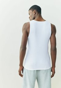 Biely bezrukávnik s rebrovanou textúrou a klasickým strihom. Nosený so sivými teplákmi, čo vytvára jednoduchý a neformálny štýl.