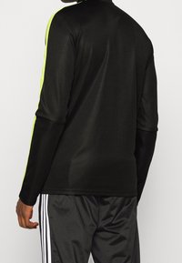 Svart långärmad träningsöverdel med subtil glans, som har neon-gula detaljer längs axlarna, kombinerad med svarta träningsbyxor.