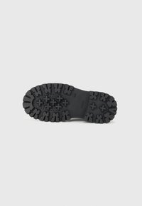 Suela de zapato de goma negra con profundas ranuras y un patrón texturizado para agarre, con elementos circulares y en forma de cruz prominentes.