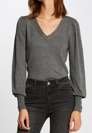 Frau trägt einen grauen V-Ausschnittpullover mit aufgepufften langen Ärmeln und geknöpften Manschetten, kombiniert mit hoch taillierten schwarzen Jeans.