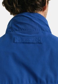 Blauwe jas met een hoge kraag, voorzien van een gestikt logo label aan de achterkant. De stof lijkt glad met een lichte glans.