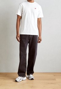 Weißes Baumwoll-T-Shirt mit einem kleinen roten Logo, kombiniert mit dunkelgrauen weiten Jeans und hellgrauen Sneakers mit silbernen Akzenten.
