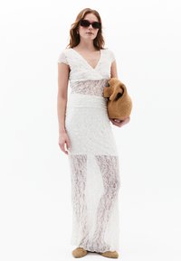 Robe en dentelle blanche avec un décolleté en V, des manches courtes et un buste ajusté. Section inférieure semi-transparente, accompagnée d'un sac tissé couleur sable.