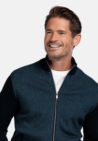 Donkerblauwe en zwarte ritsvest van textuurgebreid stof, met een hoge kraag en een zilveren rits, gedragen over een wit shirt.