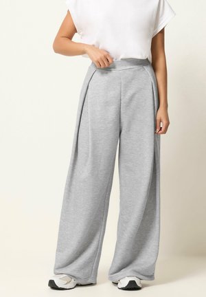 Mujer con pantalones grises de pierna ancha, camiseta blanca de manga corta y zapatillas blancas, de pie frente a un fondo claro y liso.