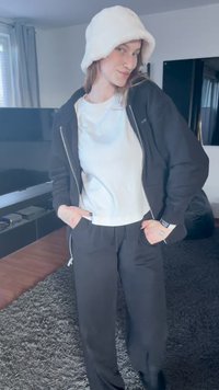 Giacca nera con zip, t-shirt azzurro chiaro e pantaloni neri. Cappello a secchiello bianco e peloso. Tessuti lisci con vestibilità casual. Sfondo scuro.