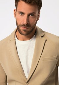 Blazer beige con un bottone, revers a punta e taschino sul petto, indossato sopra una t-shirt bianca. Tessuto dalla texture liscia.