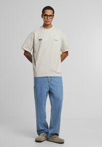 Beige oversized t-skjorte med kort erme og trykt tekst i svart og oransje. Båret sammen med lyseblå jeans og grå slip-on sko.