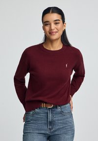 Burgunderfarbener Strickpullover mit rundem Hals und langen Ärmeln, ausgestattet mit einem kleinen gestickten Logo auf der Brust, kombiniert mit blauen Jeans.