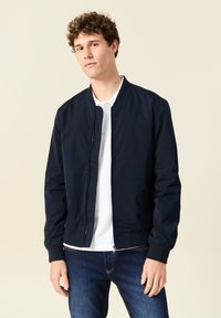 Chaqueta de piloto de la marina con cremallera frontal, puños y cuello acanalados, combinada con una camiseta polo blanca y jeans oscuros. Tejido suave y diseño sencillo.