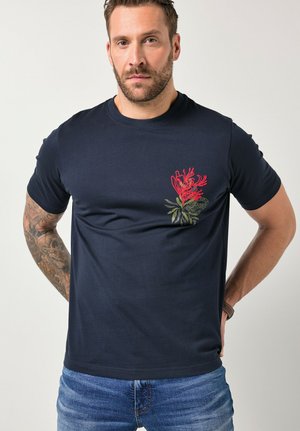 Mężczyzna o krótkich włosach i brodzie, ubrany w granatowy t-shirt z czerwonym kwiatowym wzorem na piersi oraz niebieskie dżinsy, stojący z rękami za plecami.