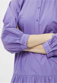 Vestido morado de botón con mangas abullonadas y puños fruncidos. Presenta un diseño en capas y una textura de tela suave.