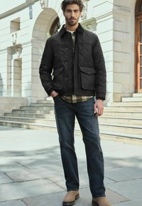 Black quilted jacket with two front pockets, worn over a plaid shirt, paired with dark jeans and tan boots. Outdoor setting with steps.