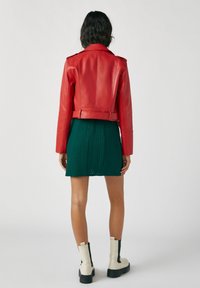 Röd läderkort jacka med axelremmar, tillsammans med en grön texturerad kjol. Har en midjebälte och svarta och vita ankelboots.