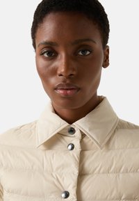Krémově zbarvená péřová bunda se strukturovaným límcem, která má přední zapínání na stříbrné knoflíky a prošívanou texturu.