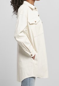 Femme aux cheveux bouclés porte une longue surchemise en velours côtelé crème avec poches et boutons, mains dans les poches latérales, associée à un jean bleu clair.