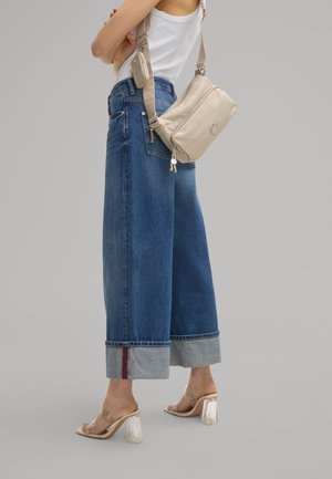 Sac bandoulière beige avec fermeture éclair, associé à un jean bleu taille haute à jambes larges avec ourlet retroussé et détail de couture rouge. Chaussures transparentes à talons blocs.